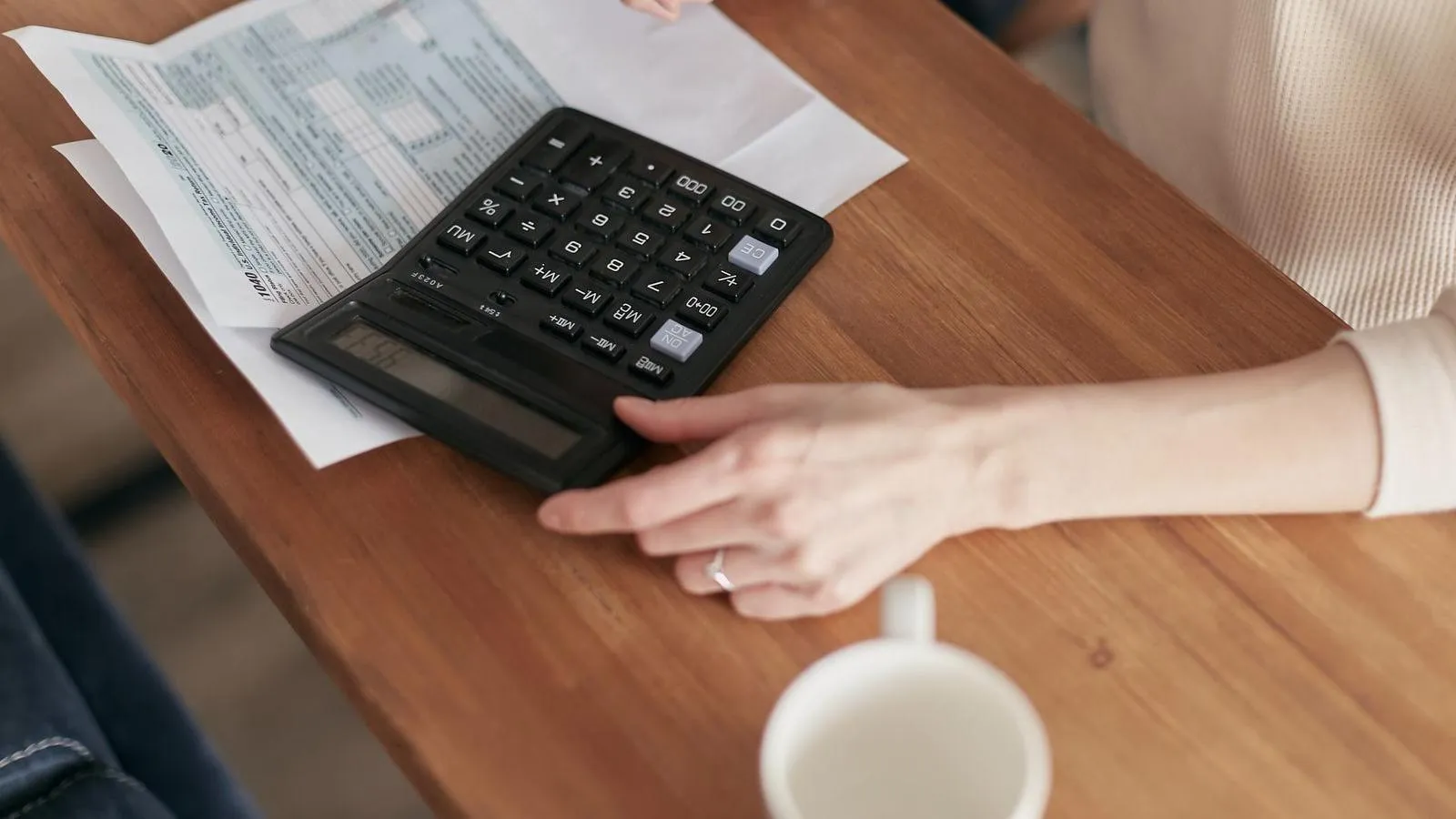 Une main posée près d'une calculatrice et de formulaires, sur une table en bois, à côté d'une tasse.