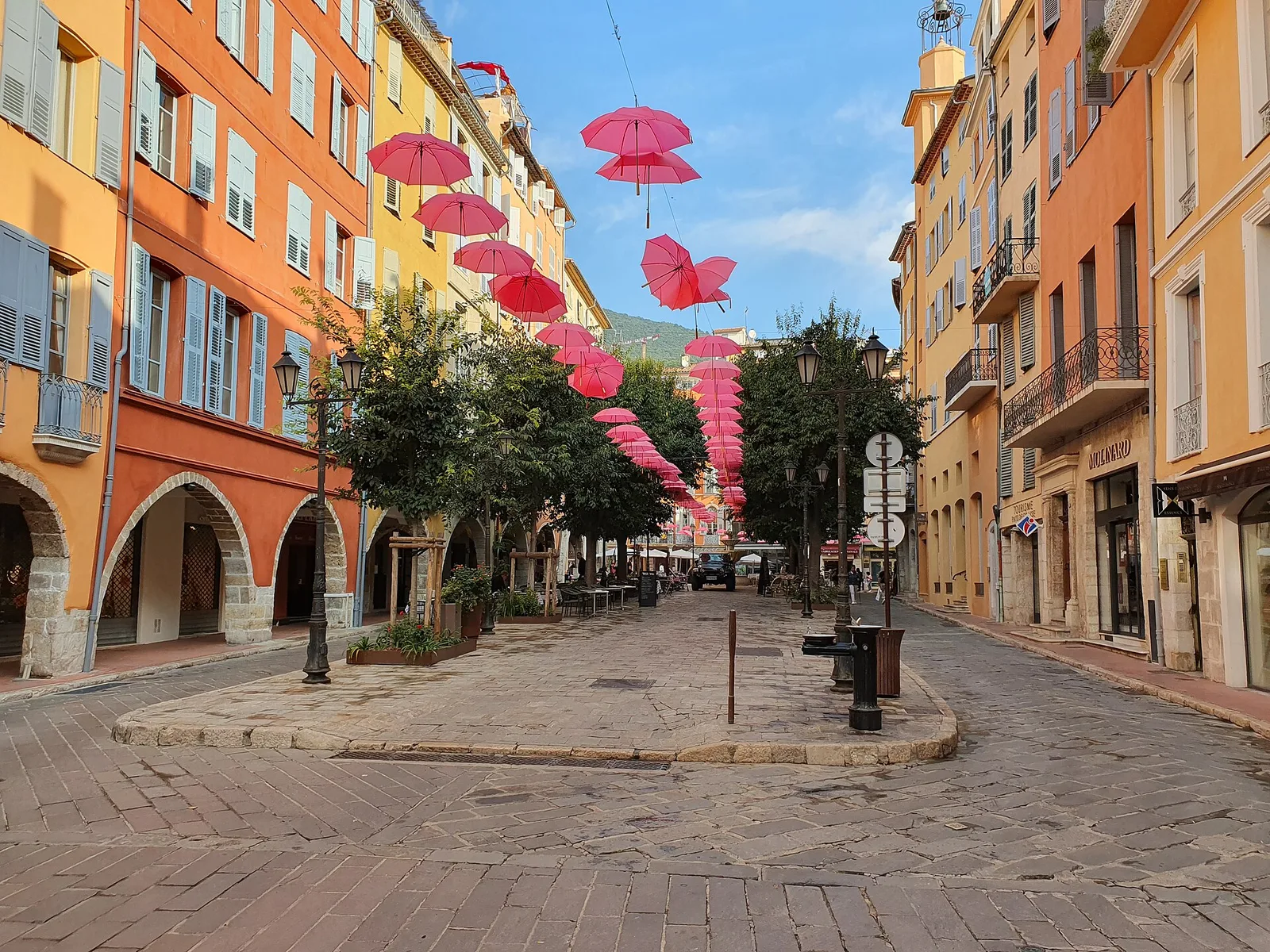 Une place pavée du vieux Grasse, bordée de façades ocre à arcades et volets, sous des parapluies roses suspendus
