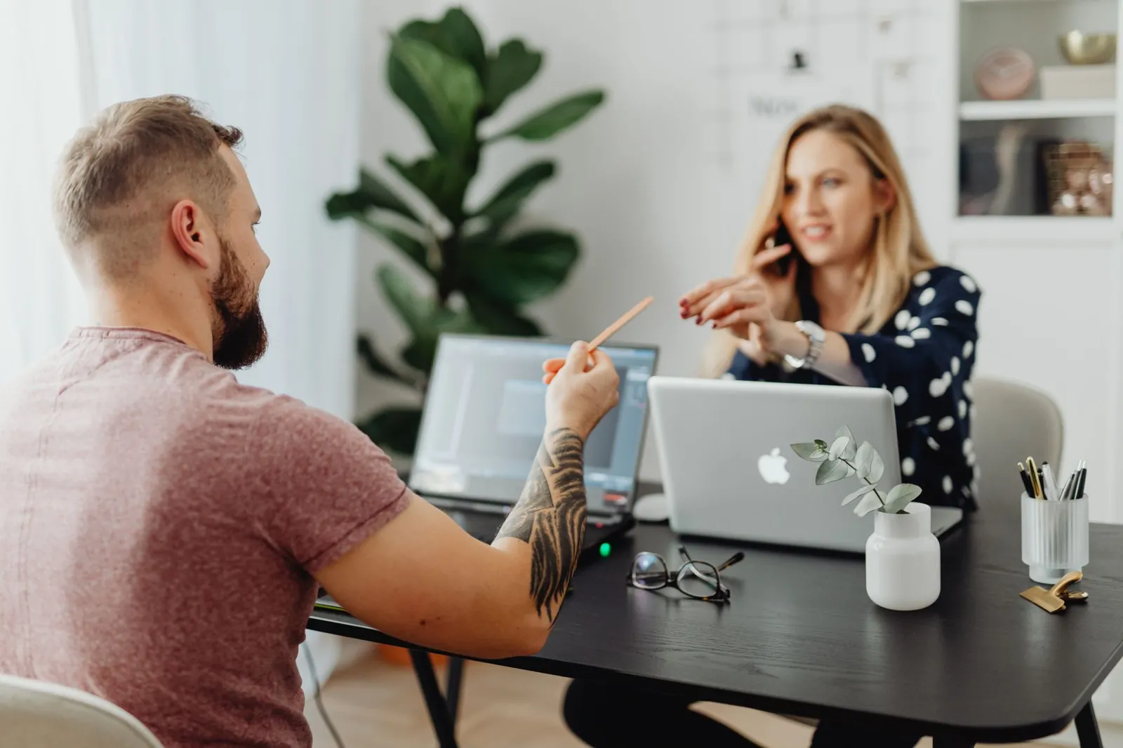Homme et femme discutant de travail à une table avec des ordinateurs portables dans un bureau moderne.